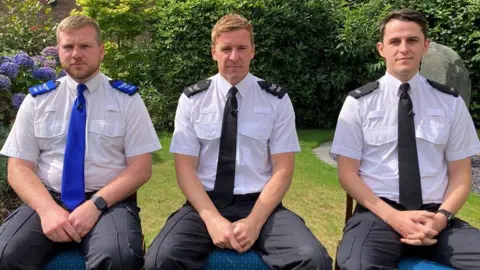 BBC PCSO Tim Parry, Sgt Greg Gillespie and PC Luke Holden are sitting down on chairs in a garden. They are all wearing their police uniform. PCSO Parry is wearing a blue tie and epaulettes on his shoulders. Sgt Gillespie and PC Holden are wearing black ties and epaulettes on their shoulders. 