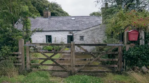 BBC News A one story white house surrounded by trees, behind a wooden gate. It has a grey roof and a red post box outside. 
