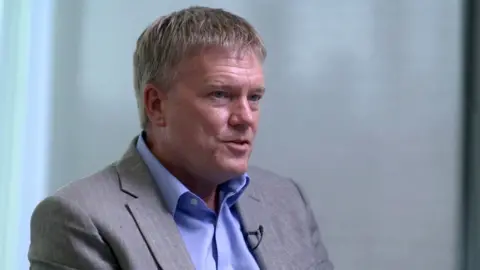 Richard Horne looking from left to right. He is wearing a light gray suit jacket over a blue dress shirt, and is seated in an indoor setting with a light-colored background