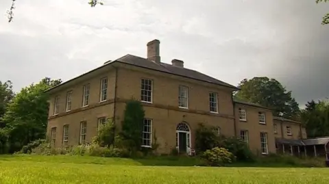 A large, two-storey country house, with a lawn in the foreground and trees behind it.