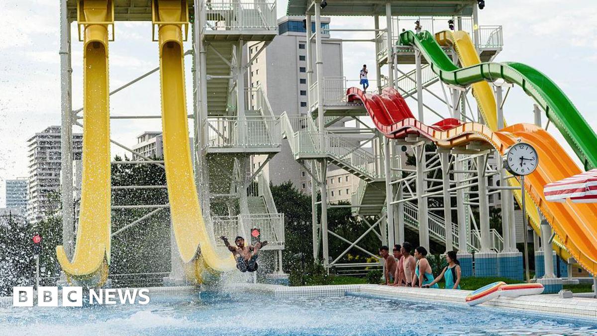 A wide shot of water slides at a water park. A man is sliding off a large yellow slide while people watch in a line beside the pool.
