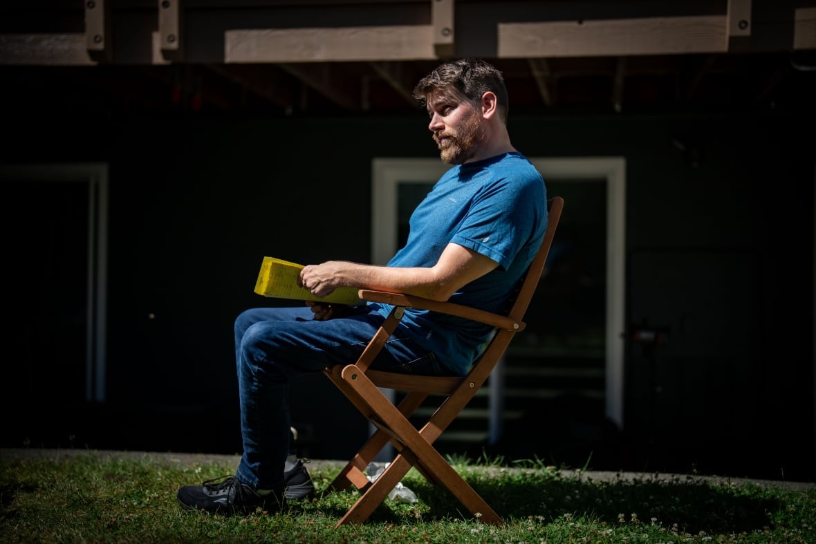 A man in a blue T-shirt sits outside in a wooden, folding chair.