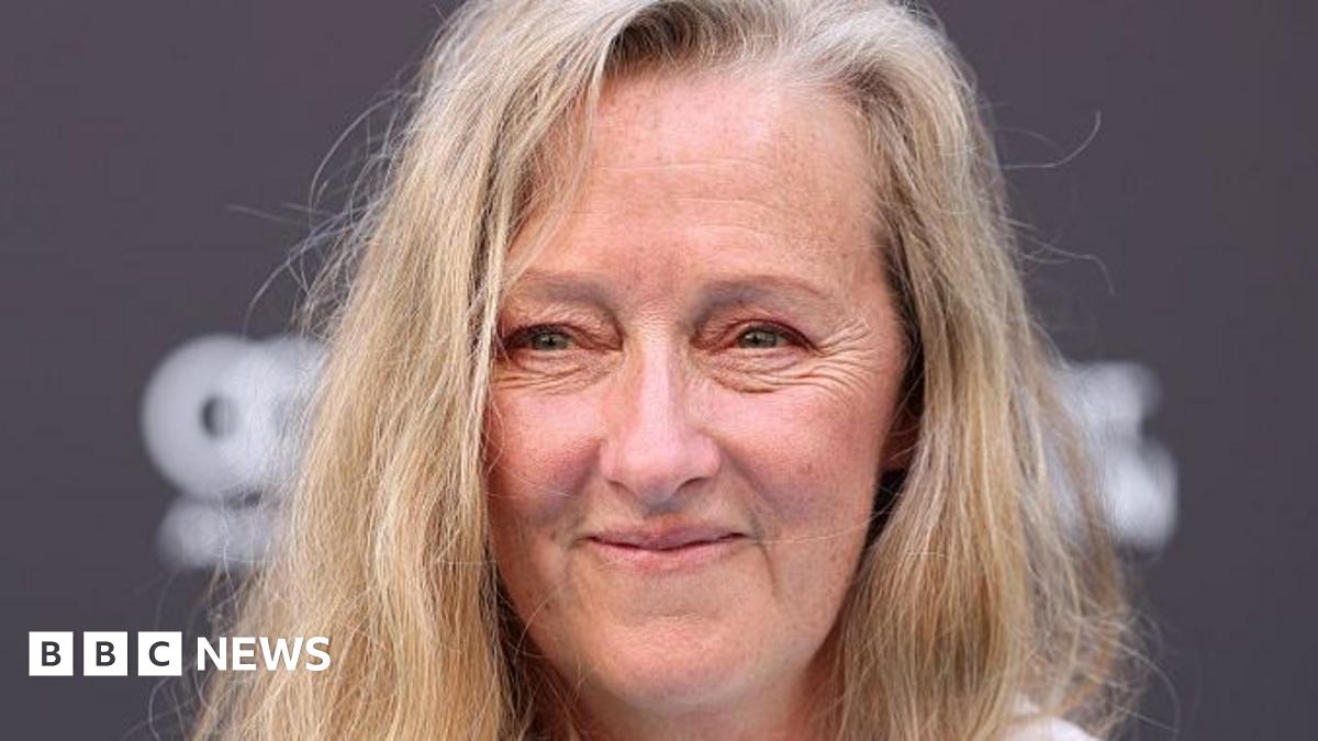 Two women stand facing the camera smiling. The women on the left has short curly ginger hair and is wearing a navy jacket. The woman on the right has short grey hair, glasses and a dark coat.