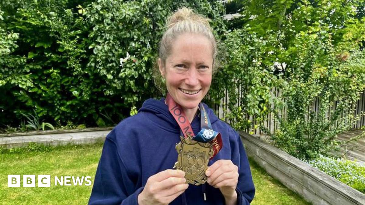 Anna Rutherford looking right at the camera and smiling. She is holding a large gold medal in front of her body. She has fair hair and is wearing a blue hooded top. She is standing in a garden surrounded by green plants and grass.