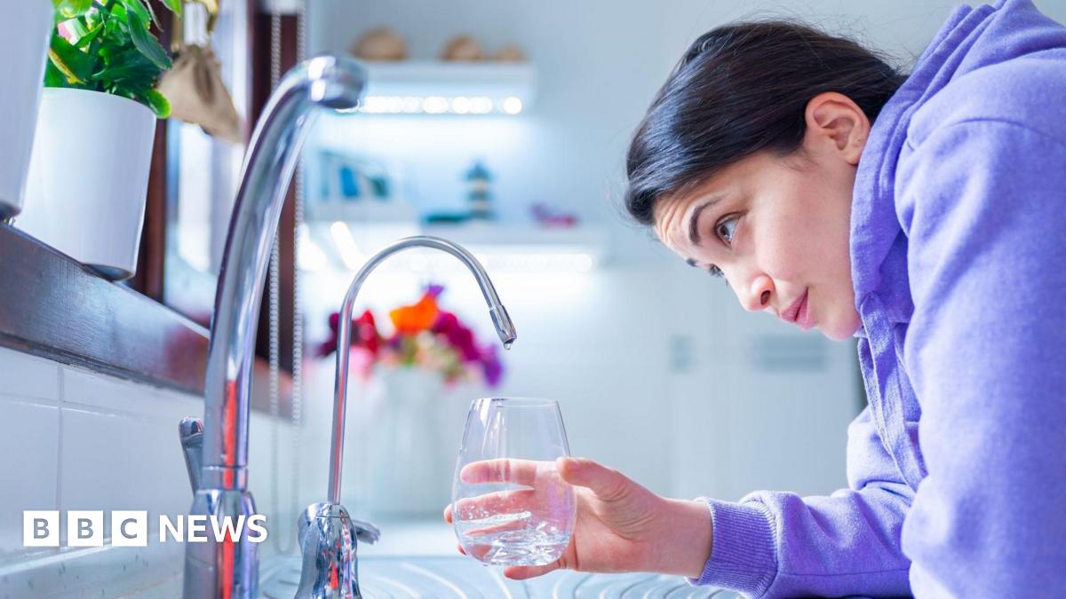 Young woman in a purple hoodie leans over a kitchen sink, holding a glass under a mixer tap. She looks slightly concerned at the lack of water flow from the tap. There is another tap in the foreground.