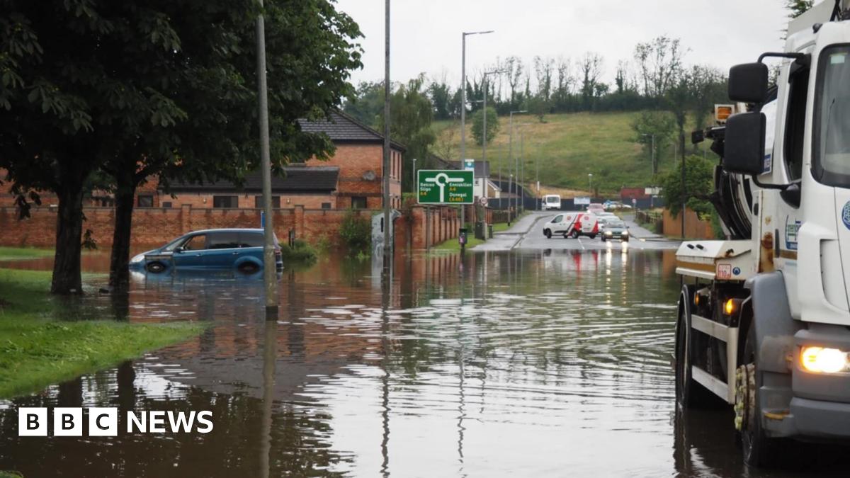 Flooding on the main Enniskillen to Dublin Road in Enniskillen on Sunday evening.  Part of the road is submerged in flood water. One blue hatchback car is parked on the left of the photo.  The water level is up to its rear bumper and more than half way up its rear wheel arch. A large white truck used to clear drains is in the foreground and cars are turning away from the area in the background.