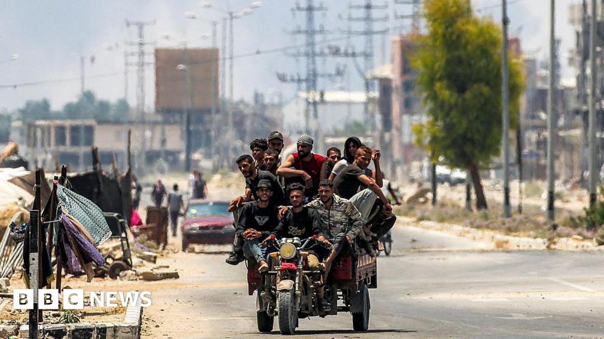Men ride on a motorised cart along Salah al-Din road in Deir al-Balah, as smoke billows in the background, in the central Gaza Strip (21 July 2025)