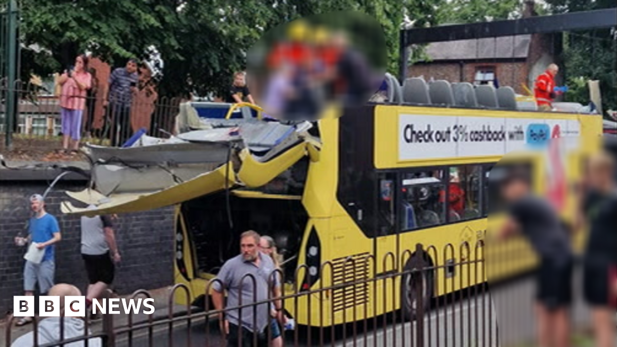 A number of people, who have been blurred out, are stood on the top floor of a double decker bus which has had its top sliced off. The back of the bus is handing off and people are watching on from either side of the road.