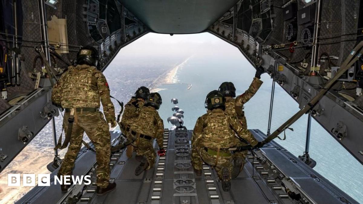 Airmen in khaki uniform watch from the open rear of an airplane as supplies are dropped by parachute