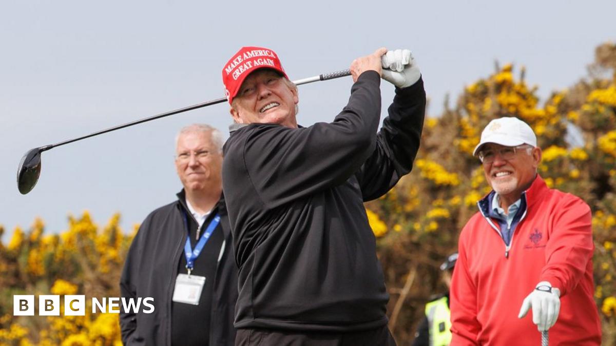 Trump on golf course wearing a red Make America Great Again cap and a white, collared golf T-shirt in May 2023. He's waving and looking directly at the camera, a green fairway behind him