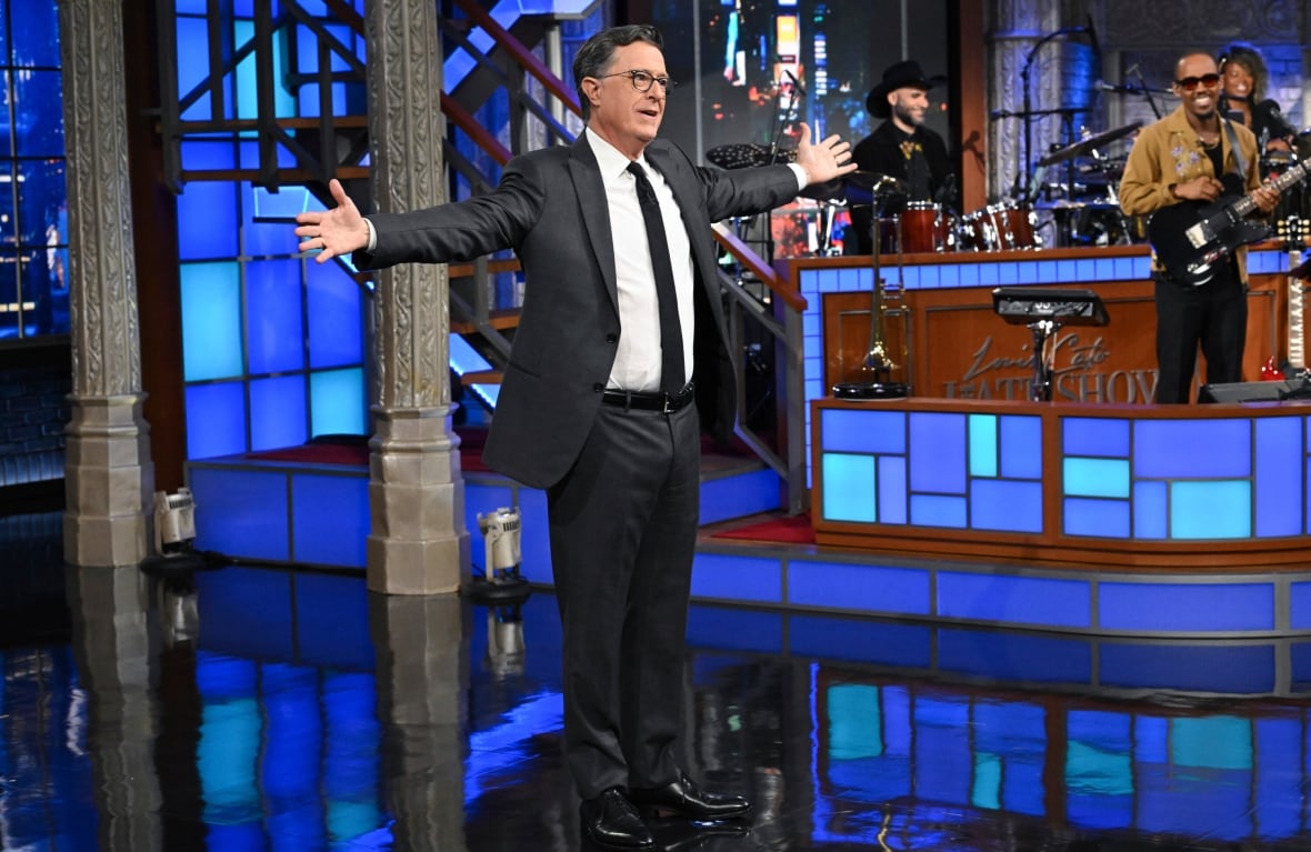 A man in a black suit, white shirt, black tie and black glasses stands with his arms stretched out on a TV set with a band in the background. 