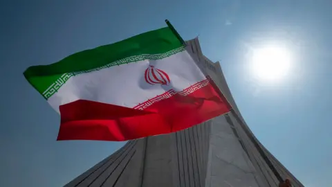 NurPhoto via Getty Images An Iranian flag is held in the air by a person out-of-frame in front of the Azadi monument against a blue sky and bright sun, in Tehran in February 2024