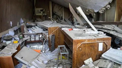 Majid Saeedi/Getty Images Damaged office equipment sits among the destruction at Evin Prison after it was hit by an Israeli air strikes. The ceiling is collapsed and piles of paperwork are strewn about. A wooden desk is in the centre of the room.