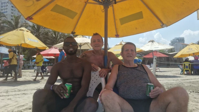 Mosquera, Alfonso and Longworth pose together under a yellow parasol on a beach. All three are smiling at the camera and holding cans of beer.