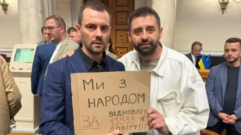 Two men, one in a suit and another in a white shirt hold a cardboard sign in Ukrainian