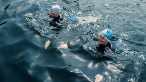 WWF Actors Miranda Richardson and Maggie Service are swimming alongside each other. They are wearing light blue swimming hats. 