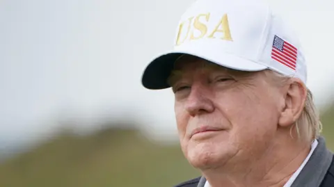 PA Media Head shot of Donald Trump smiling at the camera. He is wearing a white baseball cap with USA in gold letters and a US flag on its left-hand side.