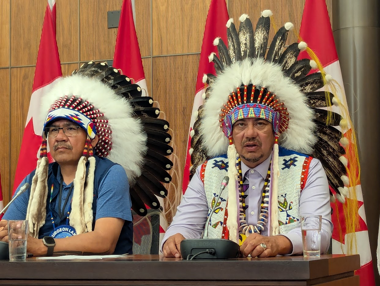 Two men in traditional garb speak in front of microphones with the Canada flag in the background.