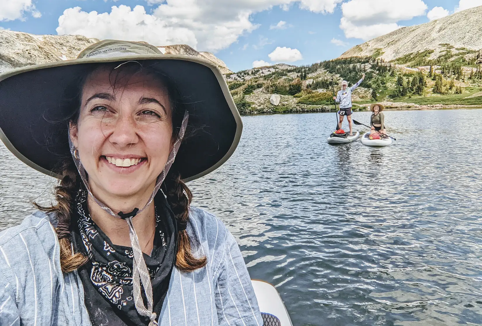 A person wearing a hat takes a selfie in front of two paddleboarders on a body of water.