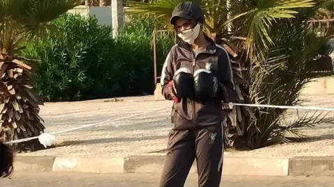 Palestinian Olympic Committee Malak Musleh standing up, wearing a black tracksuit, black boxing gloves, a black baseball cap and white facemask.