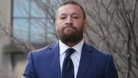 PA Media McGregor is wearing a navy suit with a white shirt and dark coloured tie. He has a thick brown beard and short brown hair. He has a straight face looking directly at the camera.