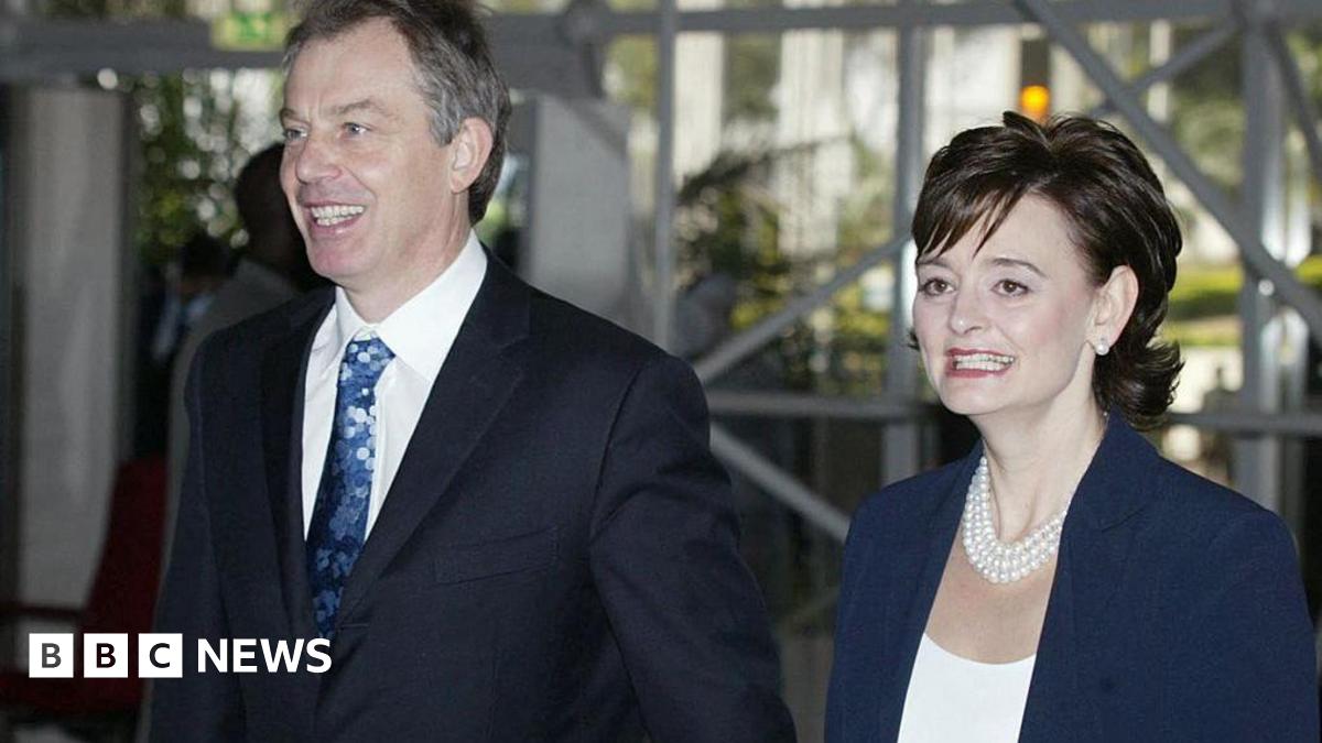 Tony and Cherie Blair walk hand in hand. They are both smiling. He has short greying hair and is wearing a dark suit, white shirt and blue patterned tie. She has medium-length dark hair and a blue jacket with a white top. She is wearing a necklace of three strands of pearls and pearl earrings.