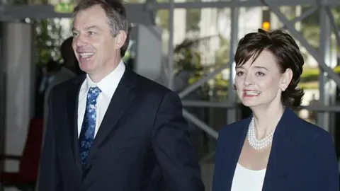 AFP PHOTO/POOL/PIUS UTOMI EKPEI via Getty Images Tony and Cherie Blair walk hand in hand. They are both smiling. He has short greying hair and is wearing a dark suit, white shirt and blue patterned tie. She has medium-length dark hair and a blue jacket with a white top. She is wearing a necklace of three strands of pearls and pearl earrings.