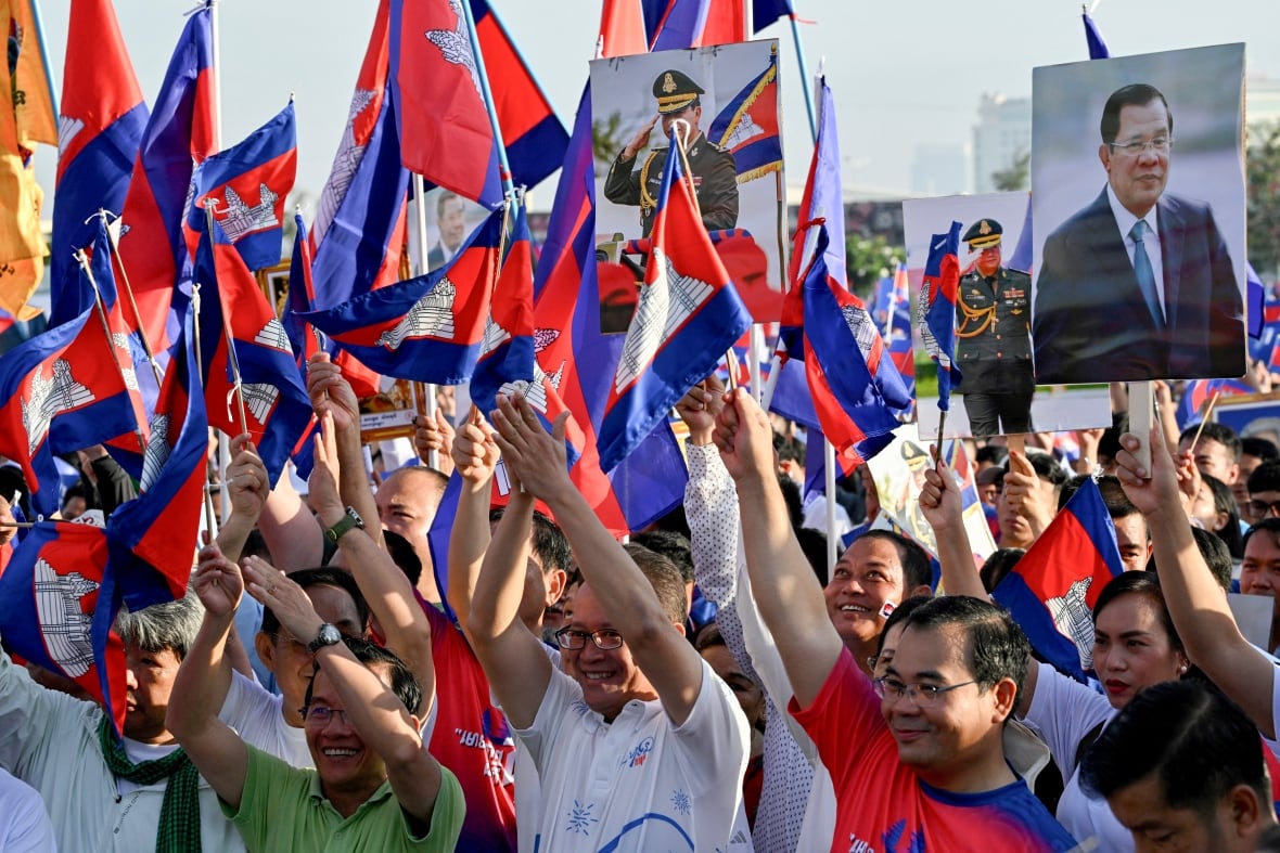 Dozens of people are shown in an outdoor demonstration, raising arms, with some holding blue-and-red flags.