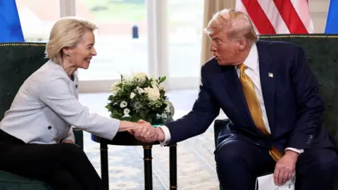 Reuters Ursula von der Leyen shaking hands with Donald Trump. They are both sitting on green chairs. Von der Leyen is wearing a grey blazer and black trousers. Trump is wearing a navy suit, white shirt and gold tie. 