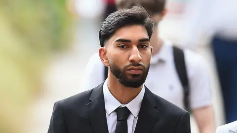 PA Media A young man wears a white shirt and a black suit and tie as he walks along a street