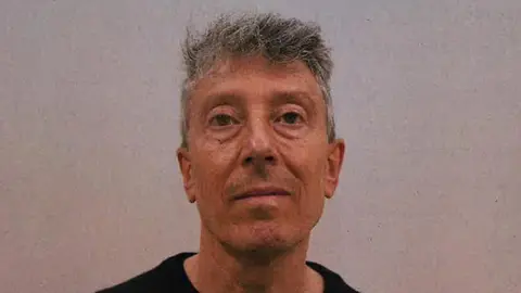 CPS A custody mugshot of Howard Phillips, who has grey hair and no expression on his face. He is wearing a black T-shirt in front of a fuzzy grey background.