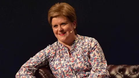 PA Media Nicola Sturgeon smiling and looking slightly off camera. She has short, orange hair and red lipstick. She is wearing a multi-coloured shirt and has her right arm slightly raised on the side of a brown sofa