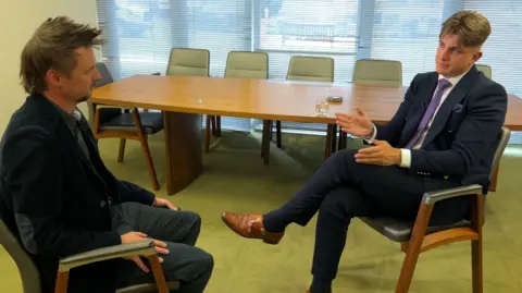 George Finch (right) speaks to BBC political reporter Simon Gilbert (left) in the Warwickshire County Council leader's office. Finch is wearing a blue suit and sits cross legged.