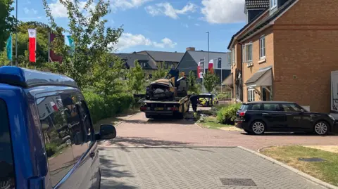 Sam Read/BBC The destroyed car is on the back of a flatbed track close to the damaged house. It is a modern estate and it is a sunny day.