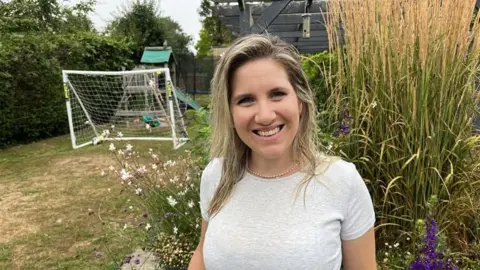 BBC/Phil Harrison A blonde woman in a marl t-shirt stands in front of a garden with a browning lawn and football goals.