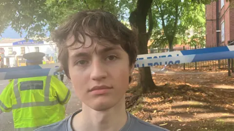 Richard Stead/BBC A young man with messy light brown hair stands with a neutral expression in front of police tape which is across an area in front of a tree. A police officer is stood facing away behind him. 