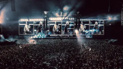 Big Brother Wide shot of Oasis on stage in Cardiff. Thousands of people can be seen standing in front of the stage. Pictures of Liam and Noel performing can be seen on the stage screens. 
