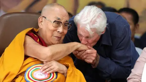EPA/Shutterstock US actor Richard Gere, in a navy shirt and with wooden bracelets on, kisses the hand of the Dalai Lama cloaked in a yellow robe with a bright multi-coloured brooch on.