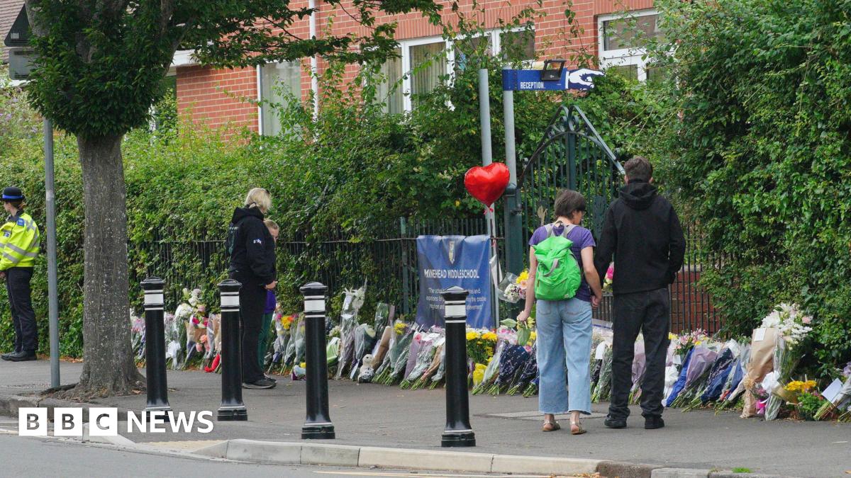 A few people look down at floral tributes placed outside Minehead Middle School. There are lots of bunches of flowers places along the pavement, next to the fence.