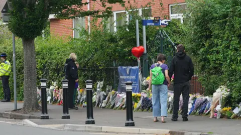 PA Media A few people look down at floral tributes placed outside Minehead Middle School. There are lots of bunches of flowers places along the pavement, next to the fence. 