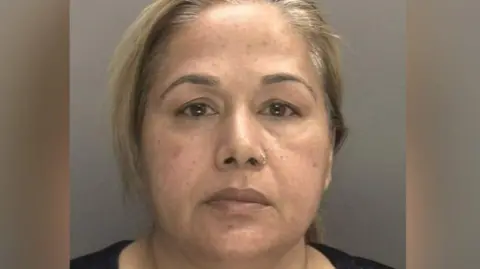 National Crime Agency Farzana Kauser with blonde hair in a ponytail and a black top, stood in front of a grey background for her mugshot.