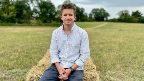 BBC Ed Caudwell smiling for the camera while sitting on a hay bale at his farm. It is a cloudy day.