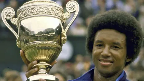 Sports Illustrated/Getty Images Arthur Ashe, in a blue tracksuit top, smiles as he holds up the Wimbledon trophy after winning the men's final on 5 July 1975.