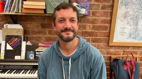 A man with short dark hair, a beard and a blue hooded top, sits and smiles in front of a keyboard pushed up against a brick wall. Two shelves behind him are filled with music-related books and other items. A framed map is hanging on the wall to the right. A black bag with red straps has been placed on a wooden chair, also to the right.