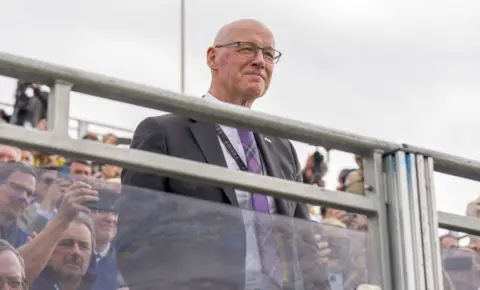 PA Media John Swinney with a grey suit and tartan tie stands with a crowd looking over a barrier