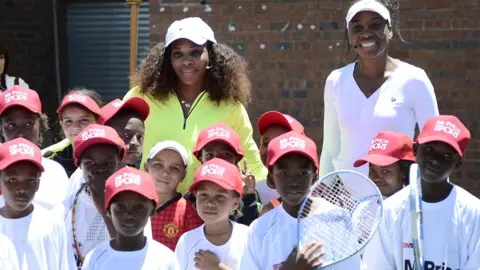 AFP/Getty Images US tennis player Serena Williams (left) in a yellow top and white cap flanked by her sister Venus in a white  top and cap pose with children in red caps and white T-shirts, some holding rackets after a two-hour tennis clinic at the Arthur Ashe Soweto Tennis Centre - November 2012.