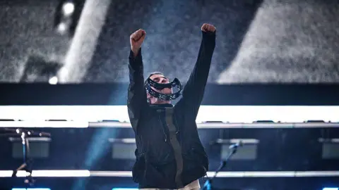 Getty Images Liam Gallagher holds a tambourine in his mouth while raising his arms in the air