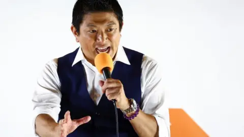 Reuters Japan's Sanseito party leader Sohei Kamiya, standing in front of his party's banner holding an orange mic, speaks to supporters on 20 July, the last day of campaigning