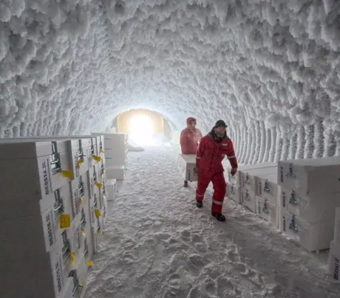 PNRA/IPEV two people in sub zero protective red jump suits carry a large white chest between them through an icy tunnel towards the camera. Lots more similar boxes are stacked on each side of the walls of the cave.