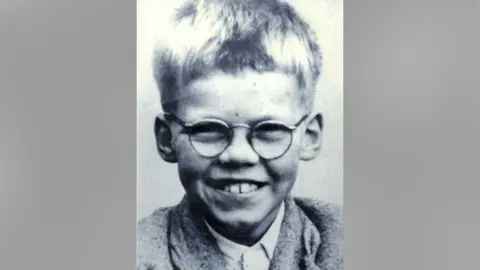Getty Images Black and white photograph of Keith Bennett with short, light-colored hair. They are dressed in a light-colored collared shirt layered under a sweater or jacket. 